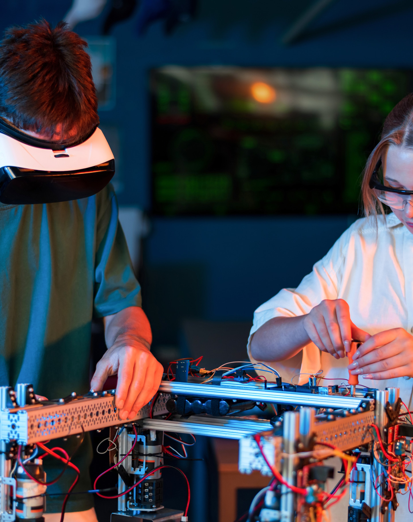 Two engineers in a lab assembling a bench-top robotics and electronics prototype, one wearing a VR headset and one in safety glasses